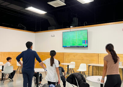 People stand around round tables in an indoor sports facility, watching a wall-mounted golf scoreboard.