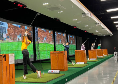 Indoor golf driving range with players at numbered bays hitting balls toward a digital backdrop and nets.
